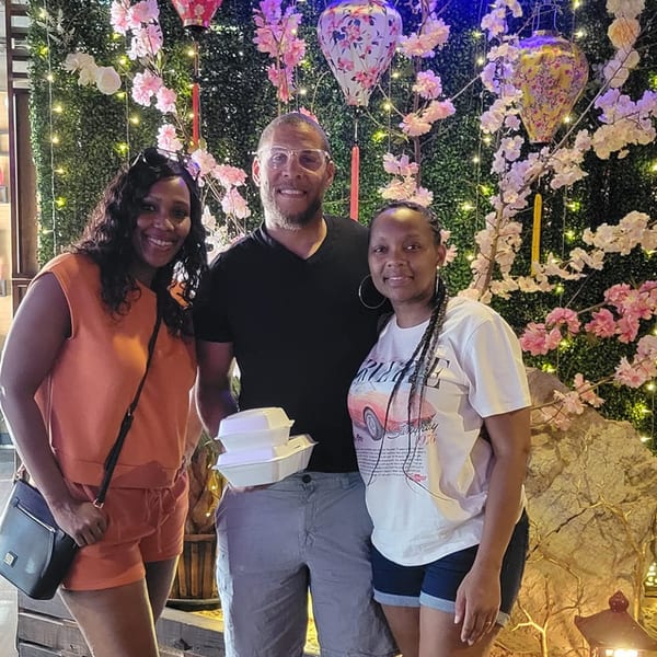 Three happy customers pose with takeout containers in front of a floral, lit-up wall at Saii Asian Bistro.