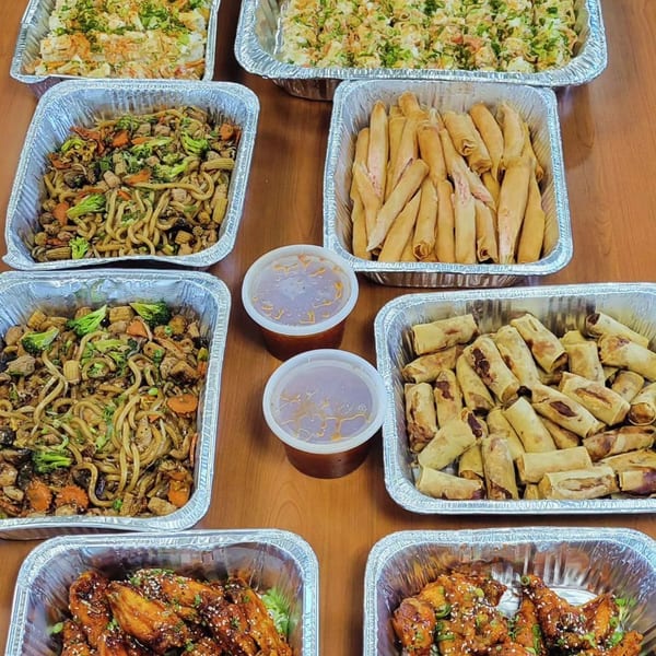 Multiple foil trays of Saii Asian Bistro's catering dishes, including noodles, spring rolls, and chicken wings.