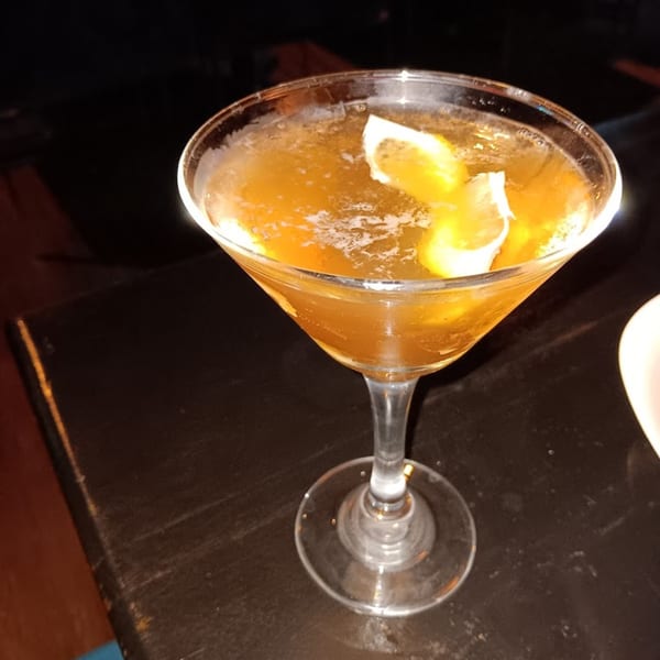 A martini glass filled with an amber cocktail and garnished with lemon peels on a dark table.