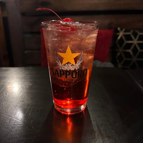 A Sapporo Premium Beer glass filled with a red cocktail, ice, and a cherry on a dark table.