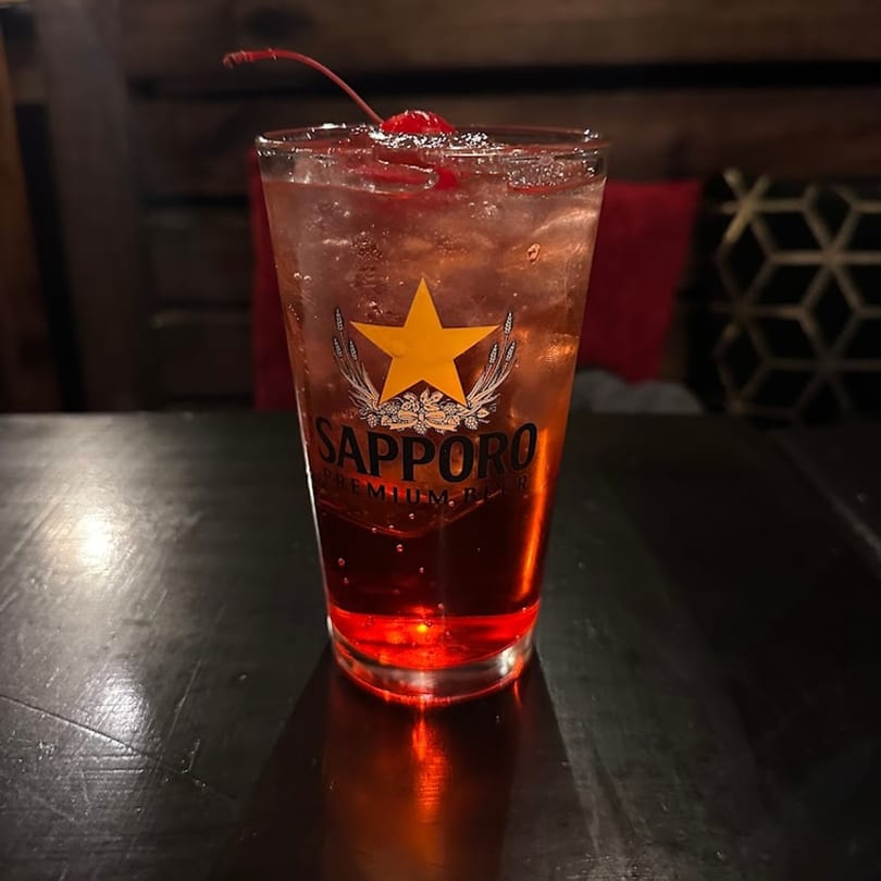 A Sapporo Premium Beer glass filled with a red cocktail, ice, and a cherry on a dark table.