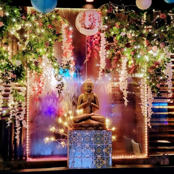 Buddha statue on a tiled pedestal in front of a waterfall, surrounded by flowers and string lights.